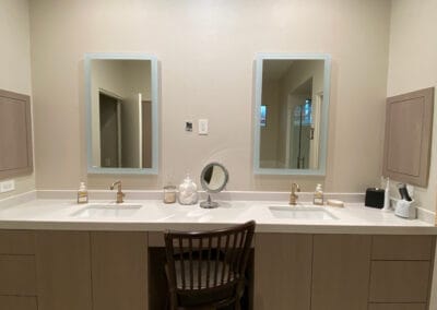 His and Her sink. Bathroom remodel featuring custom mirrors with lighting, white wood modern cabinets and gold fixtures.